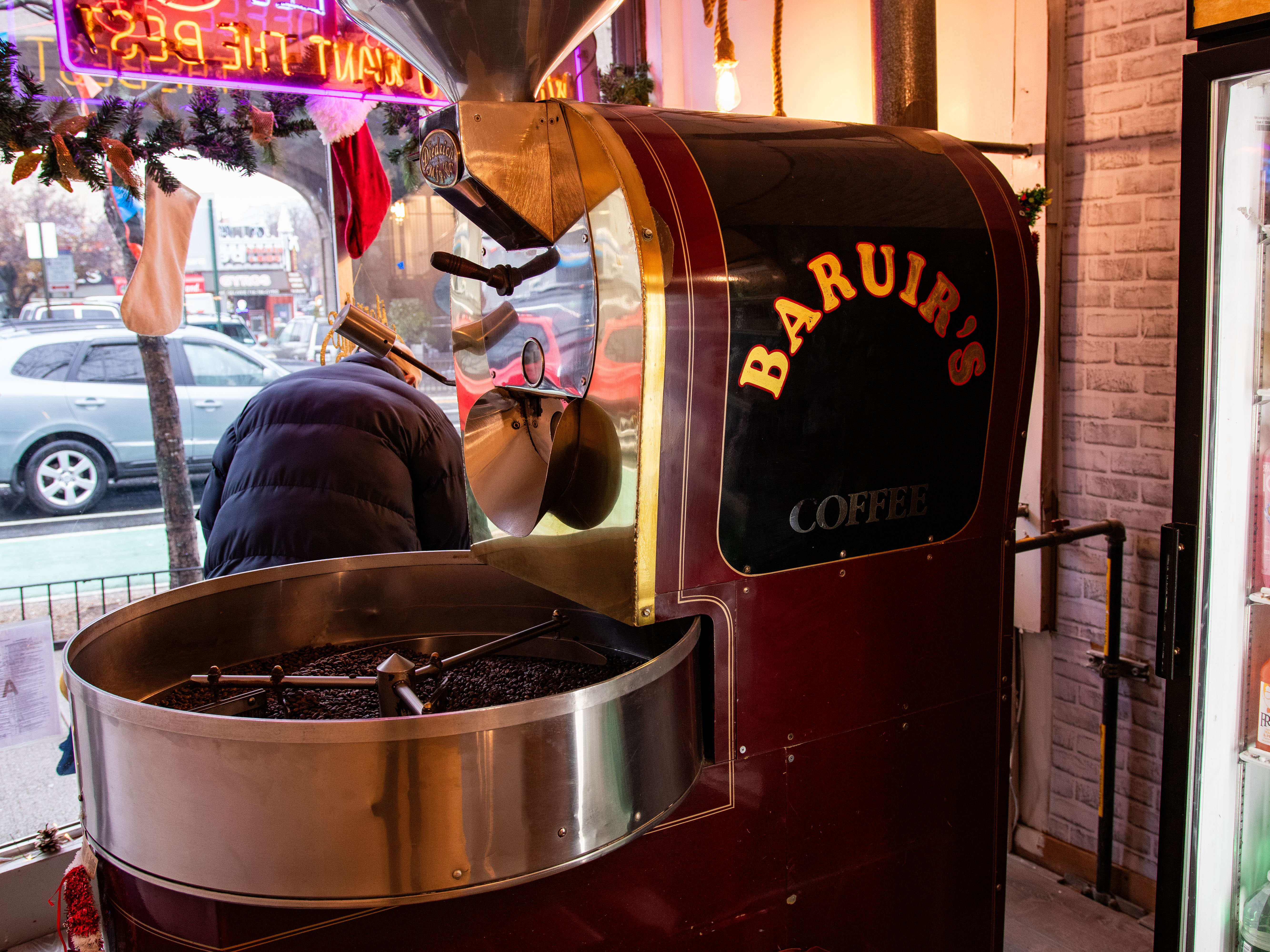 The image shows a man standing in front of the window of a café, next to a large coffee roaster with the name of the coffee shop