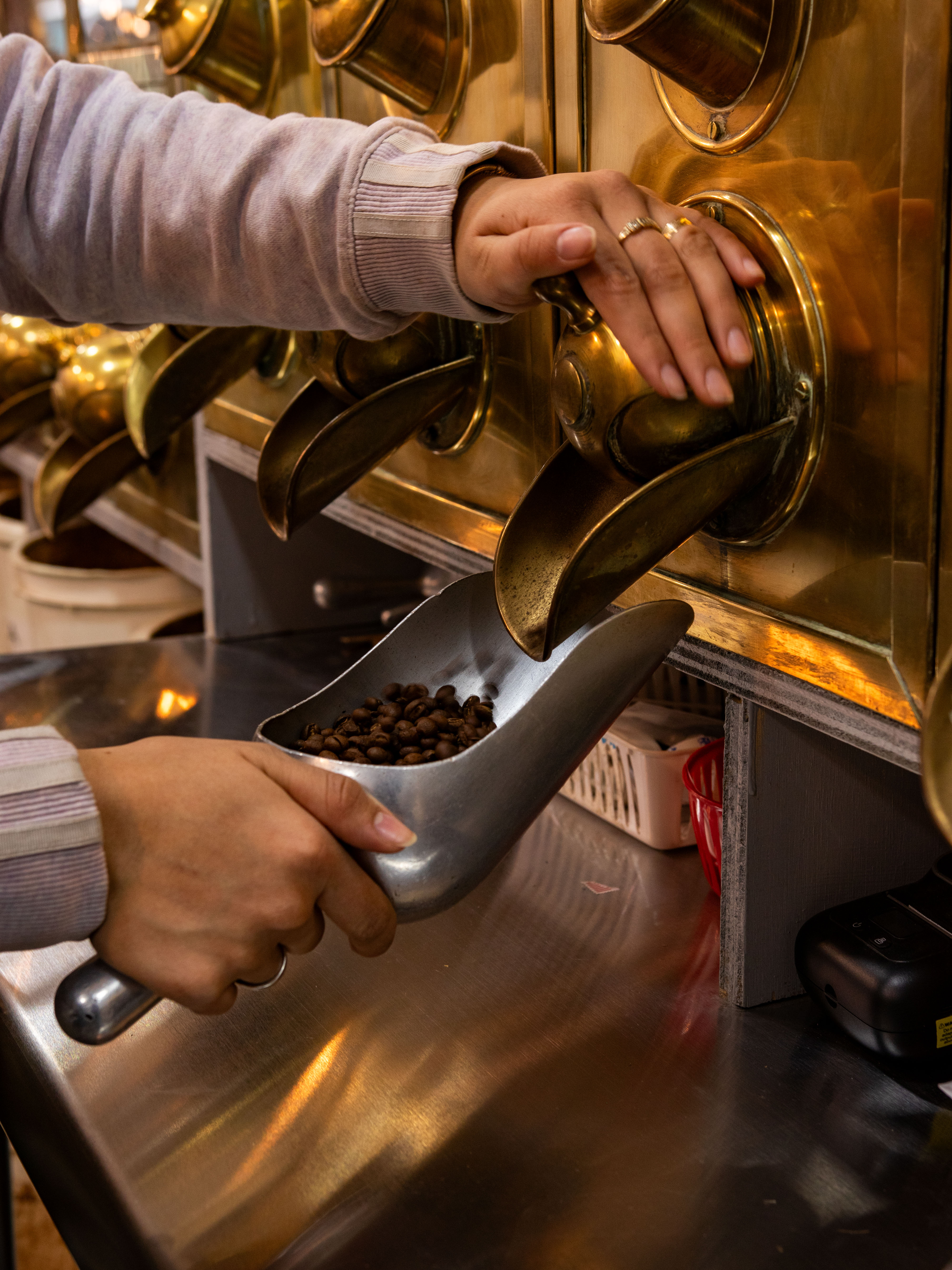 A woman's hand guiding coffee beans into a dispenser in a coffee shop or cafe, as seen in the background.